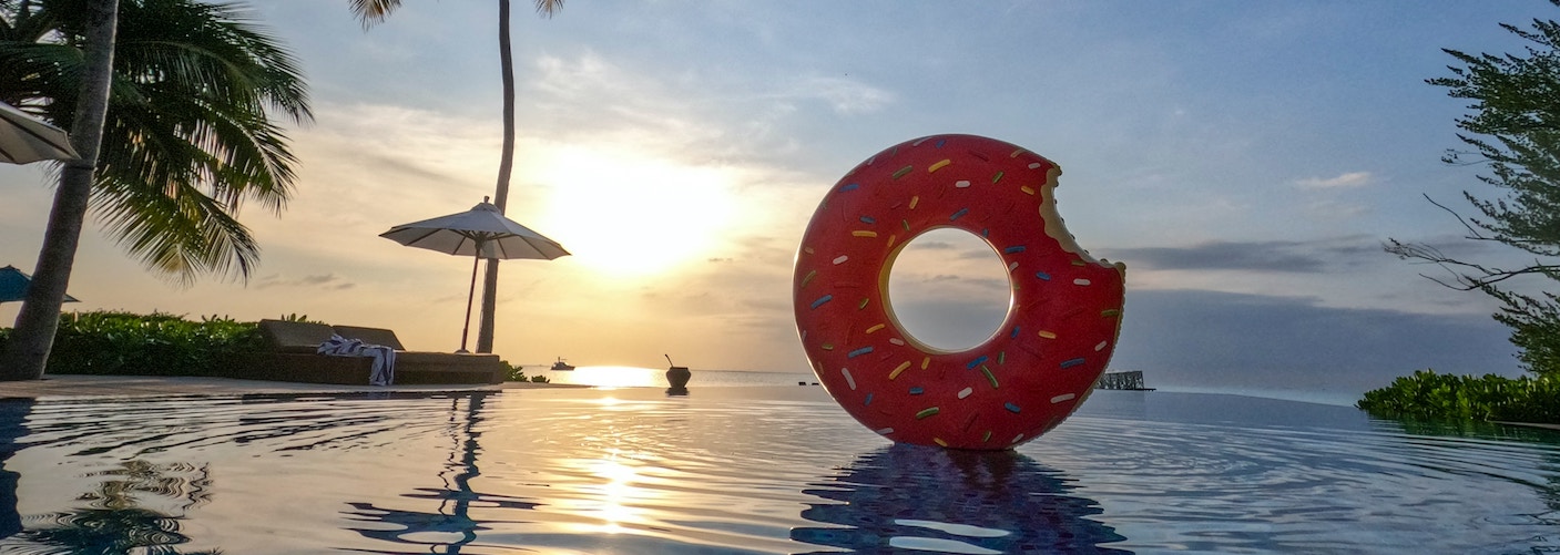 are unlimited holidays a good idea or risky business? Blog image is a big pink inflatable doughnut on the sea at sunset with a palm tree and parasol in the background.