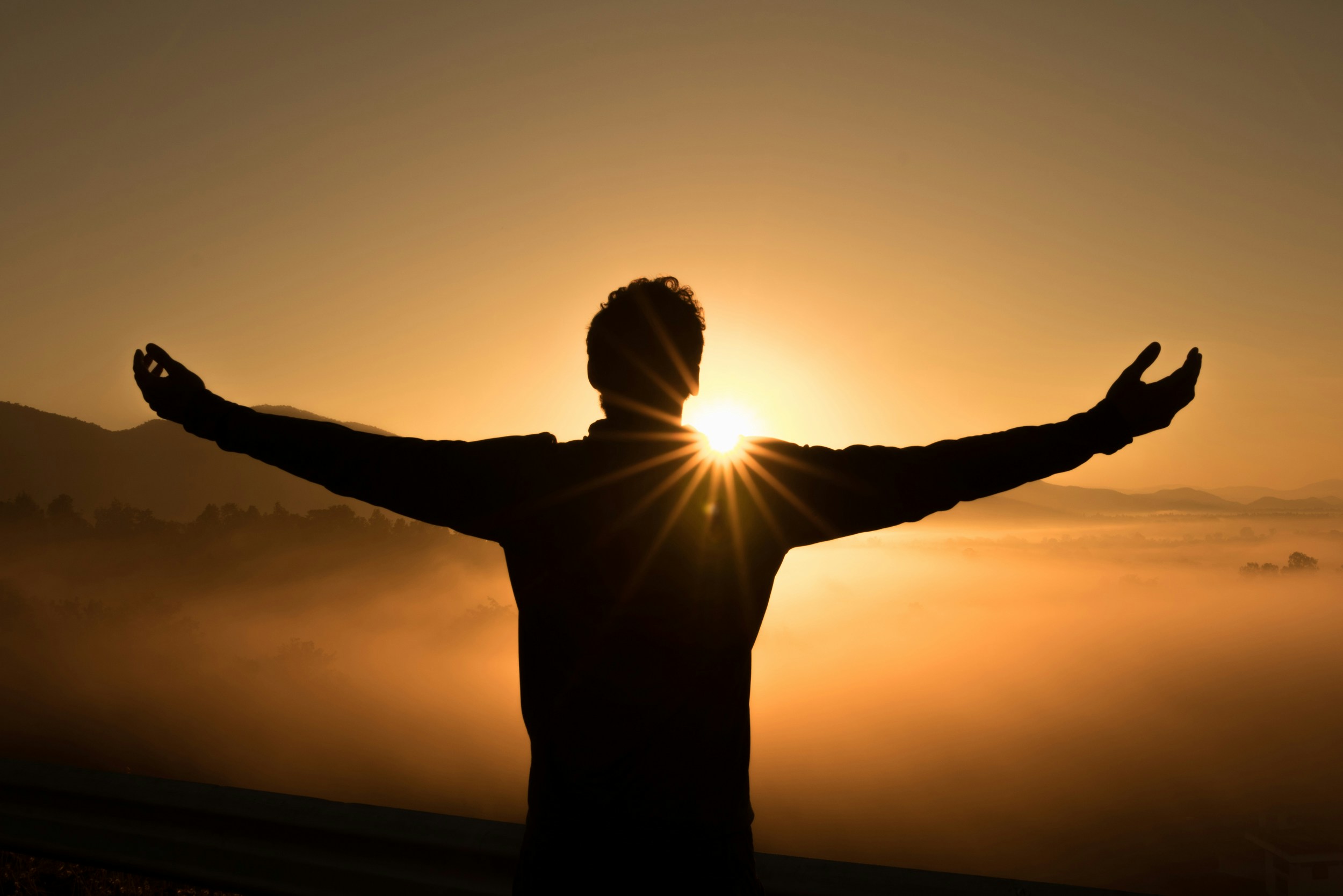 Silhouette of a person with outstretched arms facing the sunrise over a mist-covered valley.